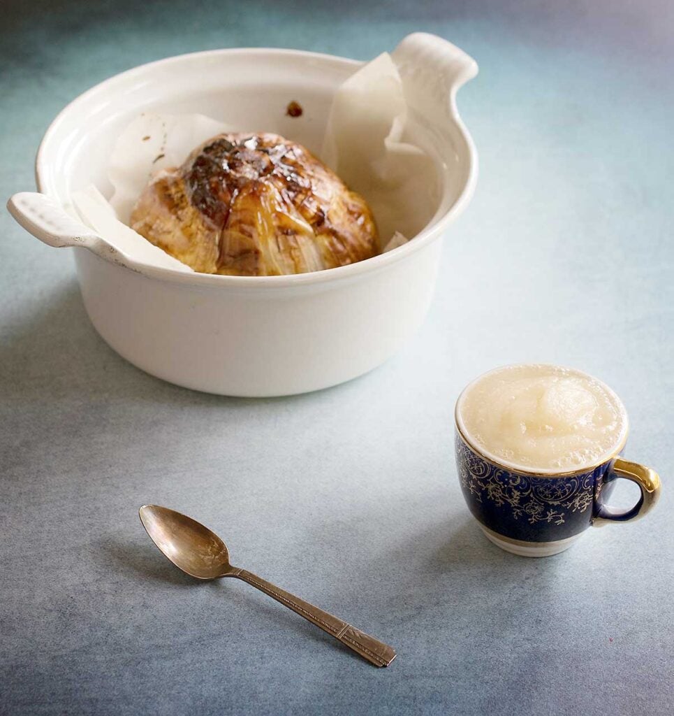 grey table with a ceramic cocotte with with a large roasted onion and a small blue and gold mug filled with onion puree dairy-free sour cream substitute.