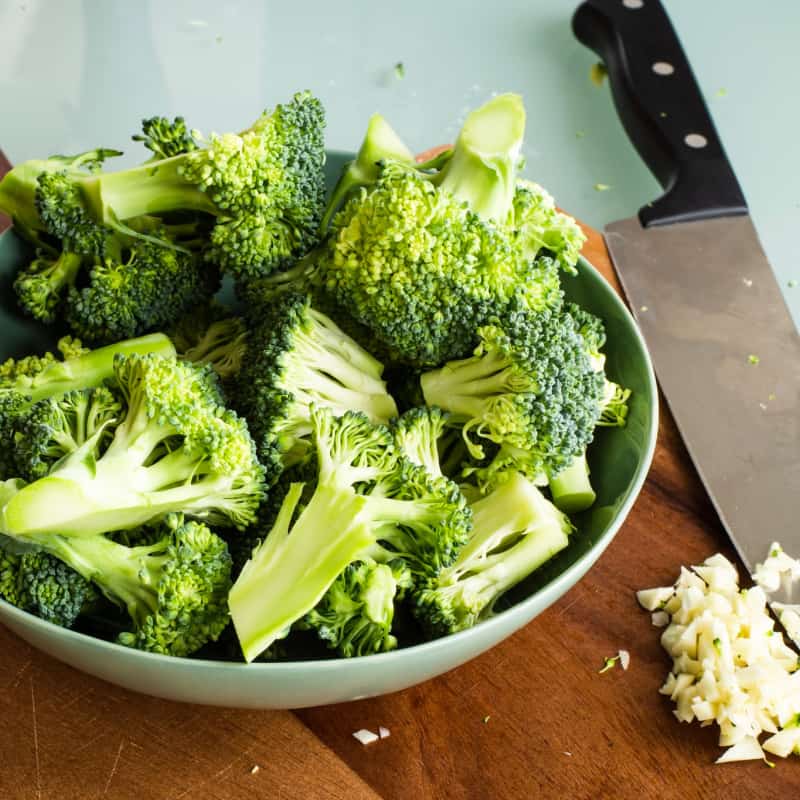 Fresh broccoli florets in a bowl on a cutting board with minced garlic
