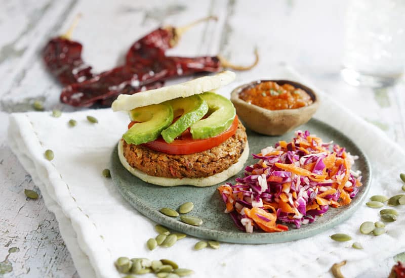 Chile Pepita Idaho® Potato Veggie Burgers with a small bowl of salsa and veggie salad in a gray serving plate with scattered pepitas and dried chilies around