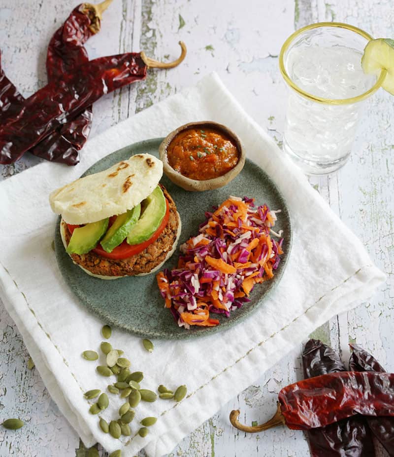 Salsa and veggie salad in a plate of Chile Pepita Idaho® Potato Veggie Burgers showing pieces of pepitas and dried chilies on a table with a glass of water