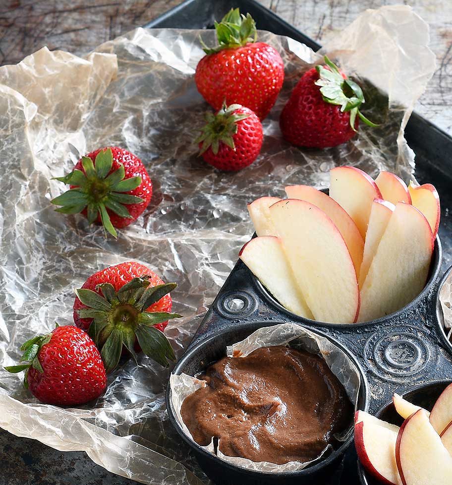 Chocolate hummus and caramel hummus in an old muffin tin with slices apples and strawberries on wax paper.