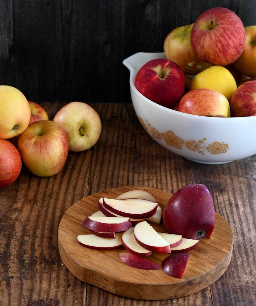 Wooden table with a Pyrex bowl of apples with an apple wooden cutting board with sliced apples on top.