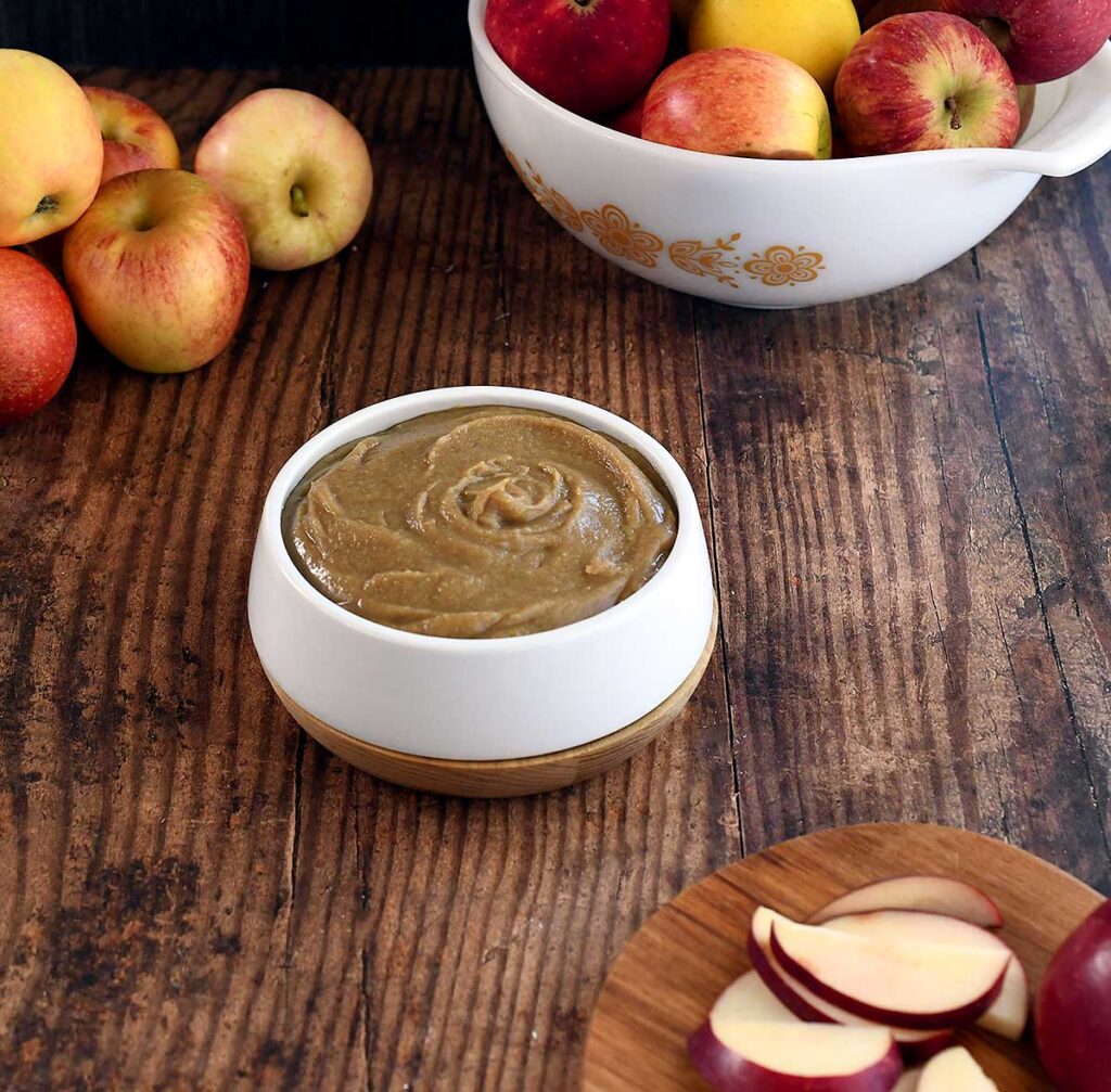 Date caramel vegan hummus swirled in a white bowl on a dark wooden tables top with apples and sliced red apples.