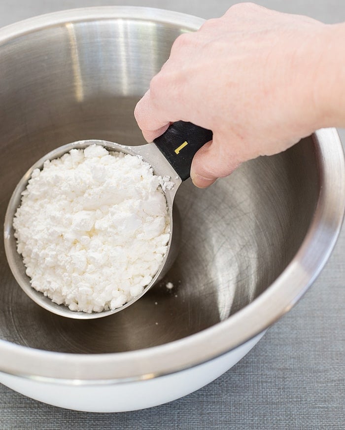 A hand holding a measuring cup filled with cornstarch ready to put in a stainless mixing bowl