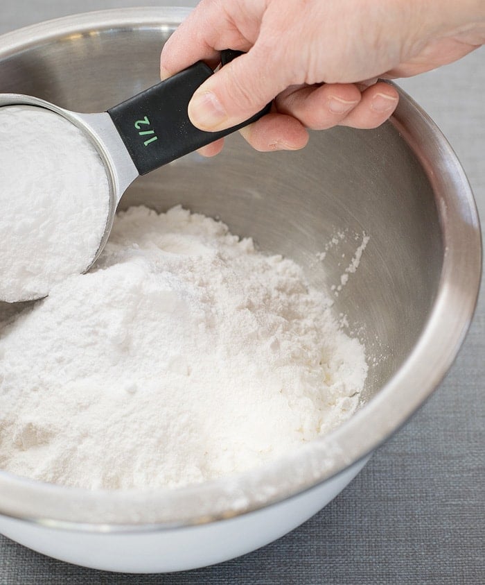 A measuring cup filled with baking soda ready to mix in with cornstarch on a stainless mixing bowl
