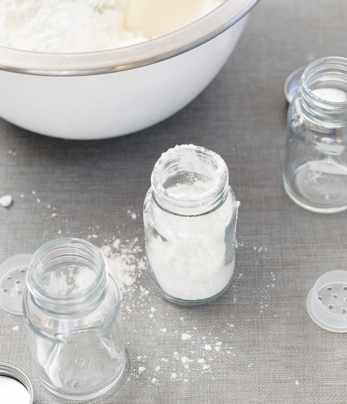 A glass bottle jar with DIY Body Powder on it and two other empty glass bottle jars beside it and scattered powder on the table