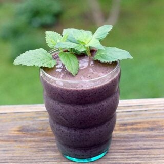 A glass full of Lemon Balm Mint Blueberry Smoothie topped with fresh mint leaves sitting on a wooden table