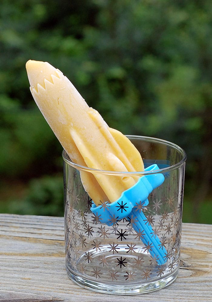 Vegan Mango Lassi Popsicles on Tovolo Rocket Ice Pop Mold Popsicle Maker inside a drinking glass sitting on a piece of wooden furniture with a shade of green leaves as a background