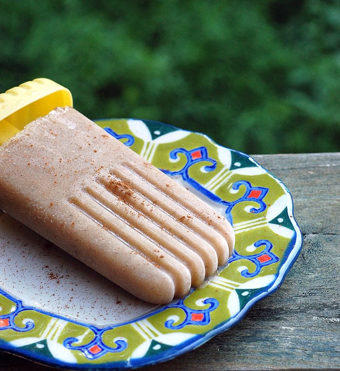vegan chai-spiced banana popsicle on a plate placed on top of wooden table outdoors, with green bush on the background