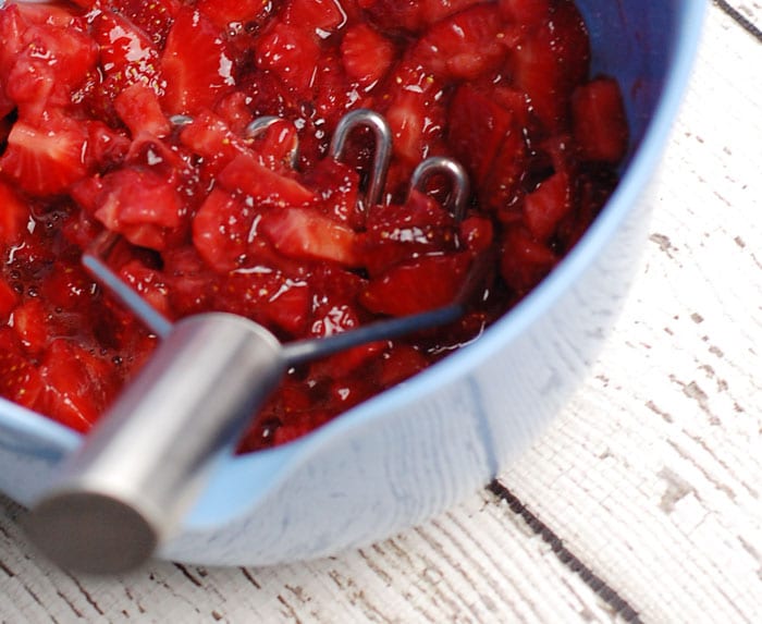 strawberries cut up and mashed in a bowl, sitting on a wooden table painted in white