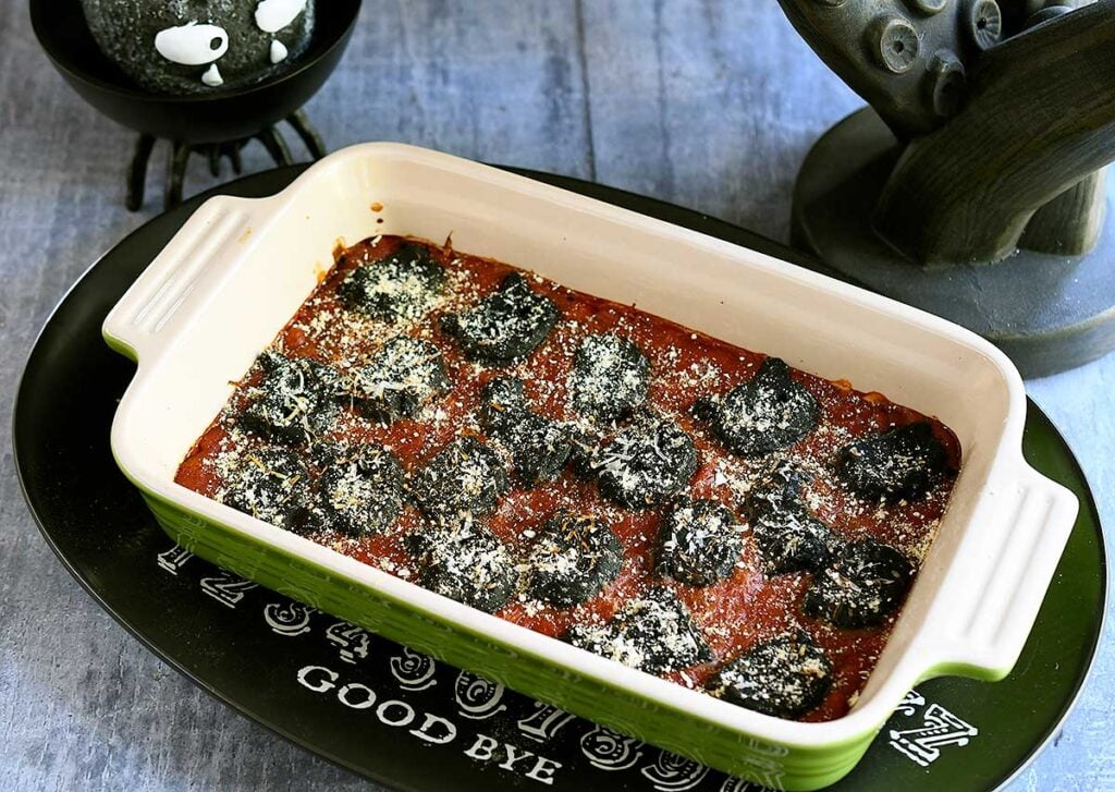 Black vegan gnocchi cut out into cat and cauldron shapes baked in a tomato sauce with spooky Halloween decorations.