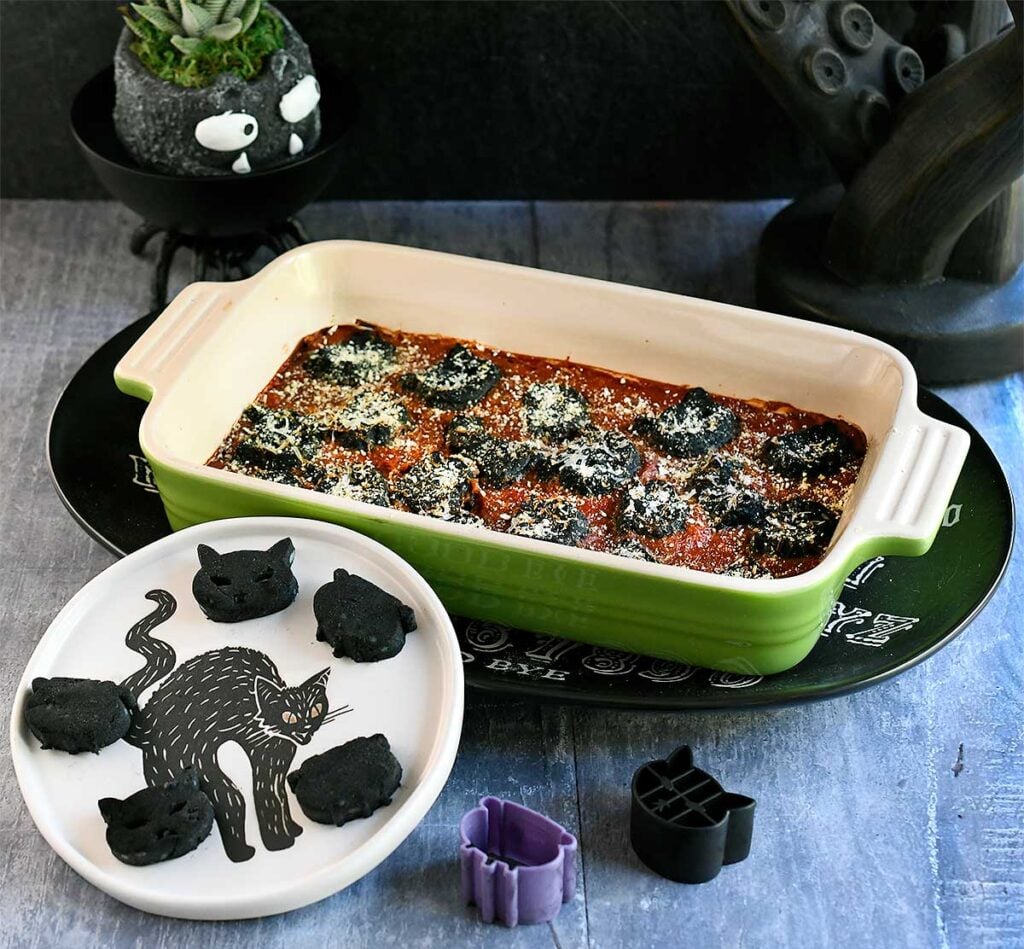 Black vegan gnocchi cut out into cat and cauldron shapes baked in a tomato sauce with spooky Halloween decorations.