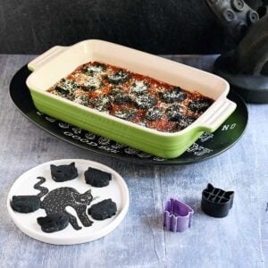 Cat and cauldron shaped black vegan gnocchi baked in a tomato sauce in a green casserole dish in a Halloween setting.