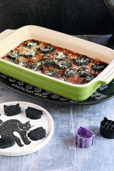 Cat and cauldron shaped black vegan gnocchi baked in a tomato sauce in a green casserole dish in a Halloween setting.