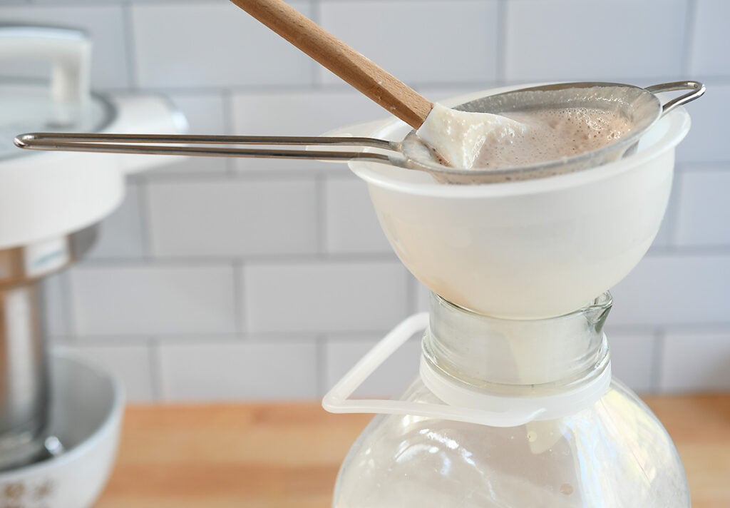 A plastic funnel in a milk jar with a bar strainer and a spatula in it used to filter the pulp.