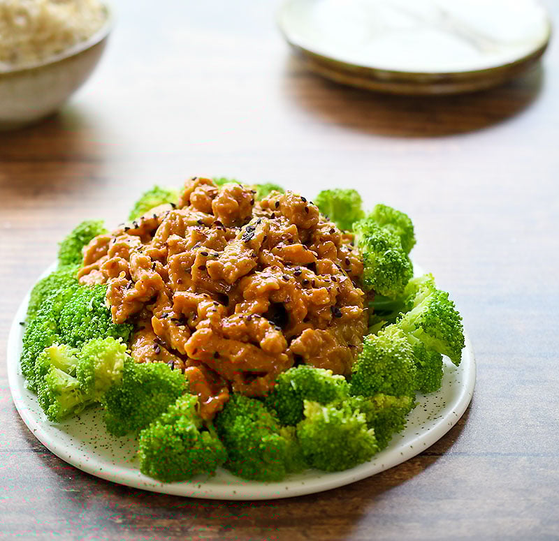 General Tso Soy Curls surrounded by a circle of steamed broccoli on a white ceramic plate on a dark wooden table