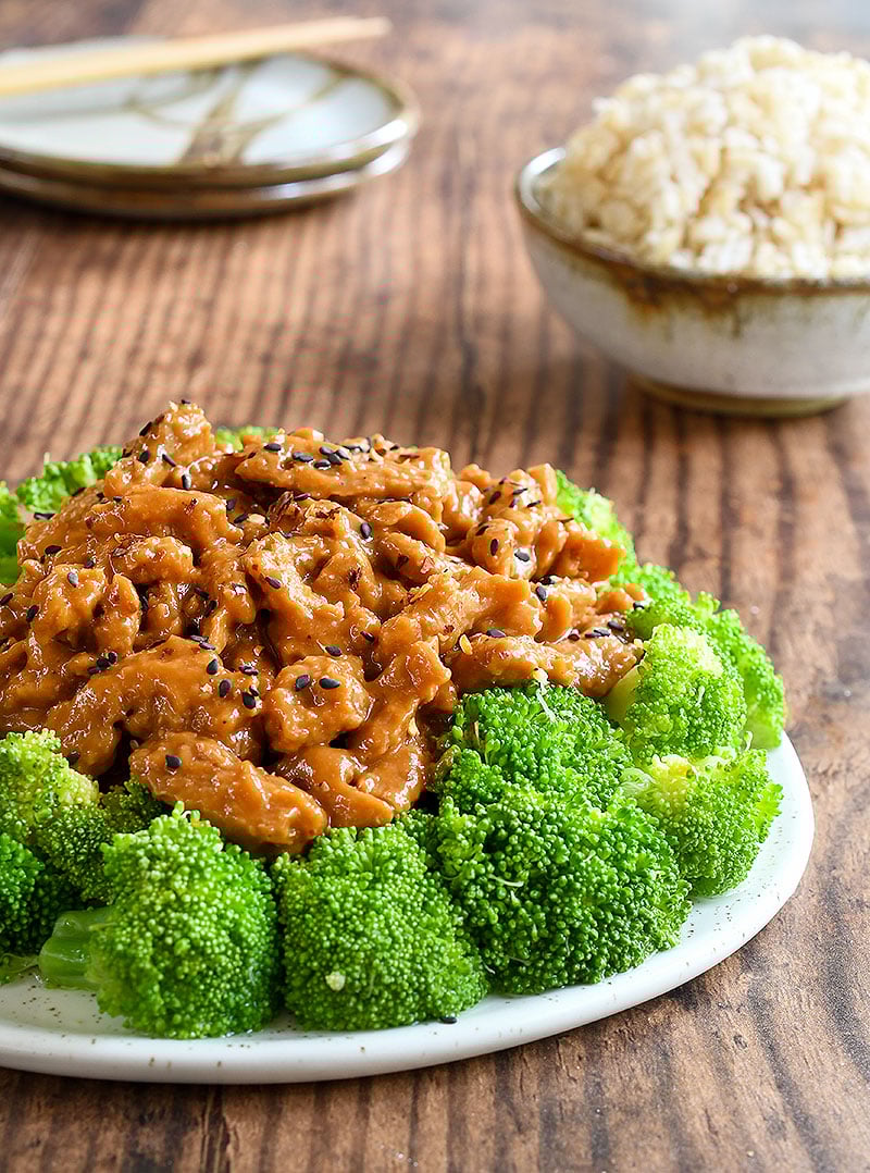 General Tso Soy Curls topped with black sesame seeds surrounded by broccoli on a white plate with a bowl of brown rice on a wooden table