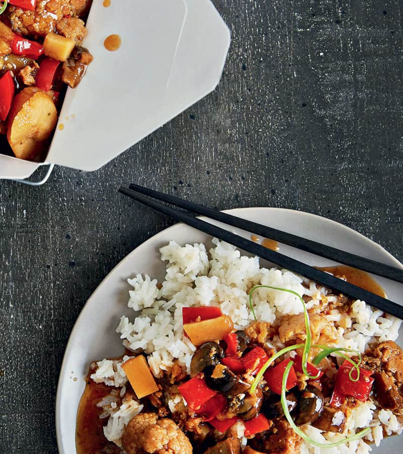 Slow Cooker Kung Pao Cauliflower served on top of a plate of white rice with chopsticks and a box of the dish on the side placed on gray matte linen