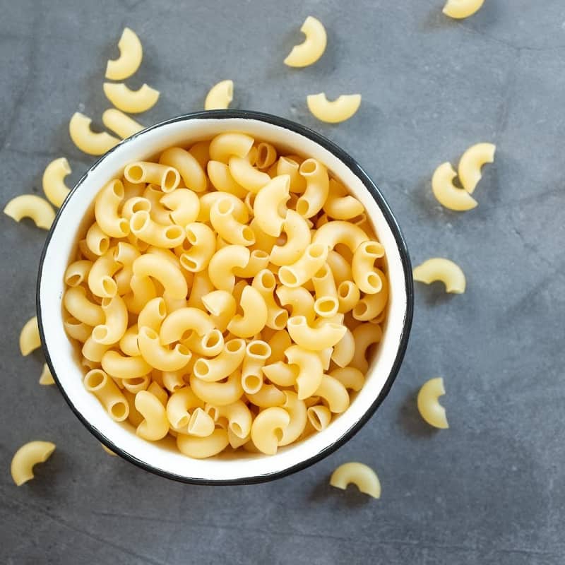 Raw elbow macaroni in enamel bowl on dark cement table background.