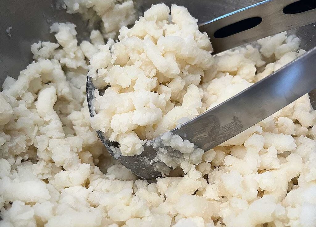 Cooked potatoes being mashed with a large silver potato masher.