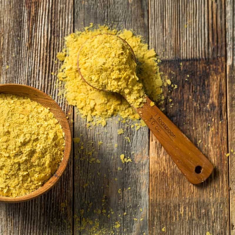 Large flake nutritional yeast in wooden bowl, overflowing in wooden spoon over planks