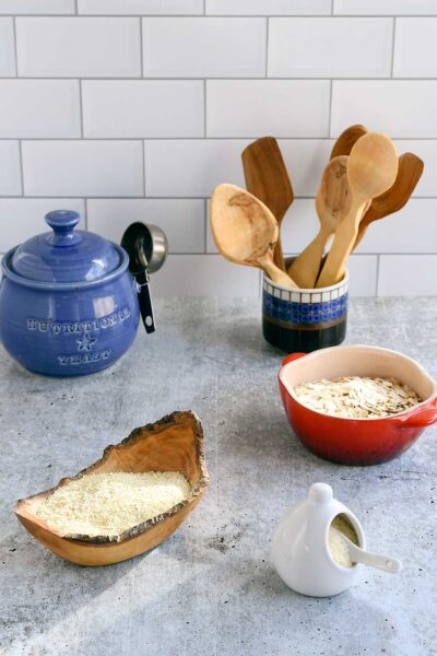 Homemade vegan parmesan in a wooden bowl, with a red ceramic bowl of oats and hand carved wooden spoons.