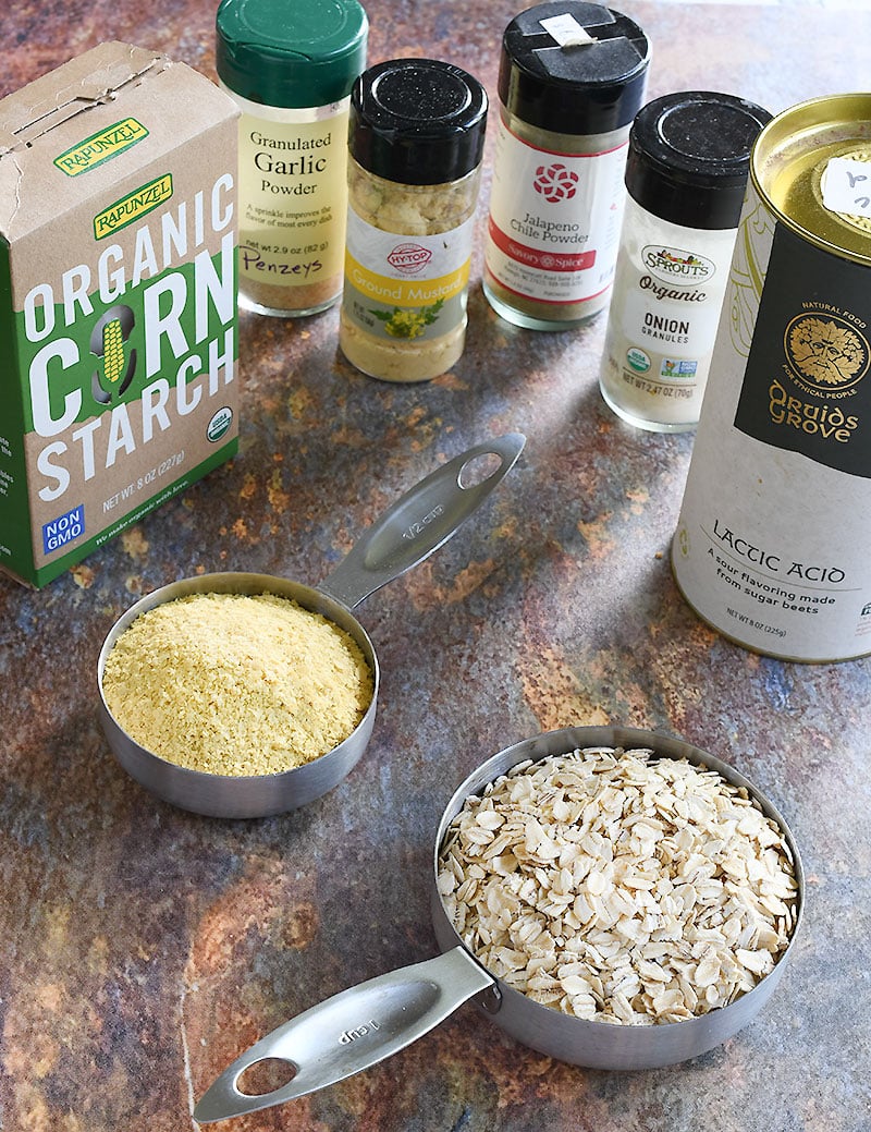 Nutritional yeast and oats on a stainless steel measuring cups sitting on a marbled table with a box of organic corn starch and mini-bottles of spices around