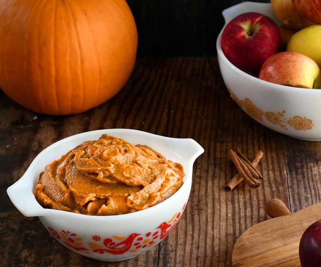 Pumpkin pie dip in a vintage white pyrex dish with red birds on it on a wooden table with a bowl of apples.