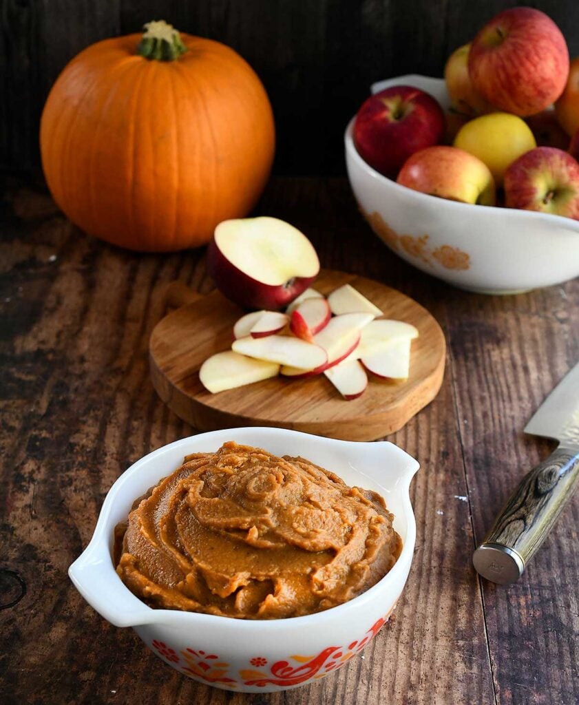 Pumpkin pie dip in a vintage white pyrex dish with red birds on it on a wooden table with apple slices and a bowl of whole red and yellow apples.