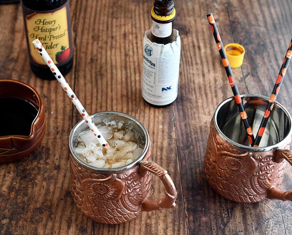 Pumpkin spice cocktail with rum in a bronze owl mug with a bottle of apple cider and bitters.
