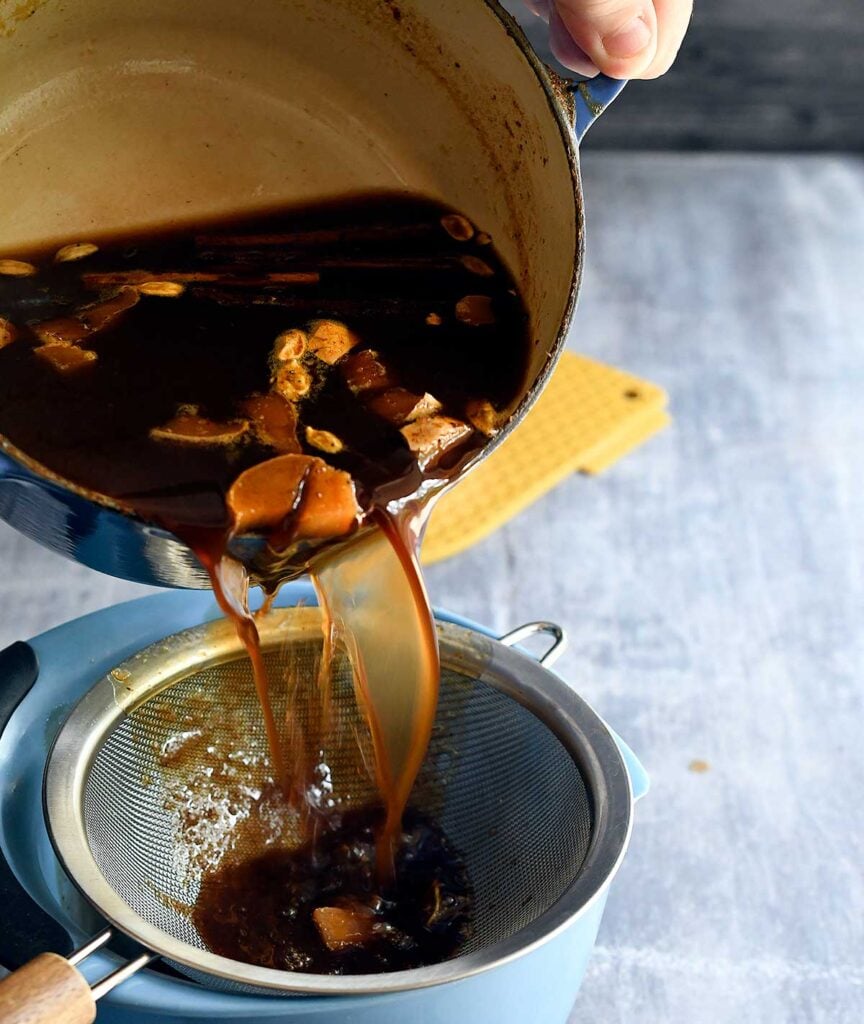 Cooked pumpkin spice syrup with chunks of ginger and spices being poured through a fine mesh strainer into a blue bowl.