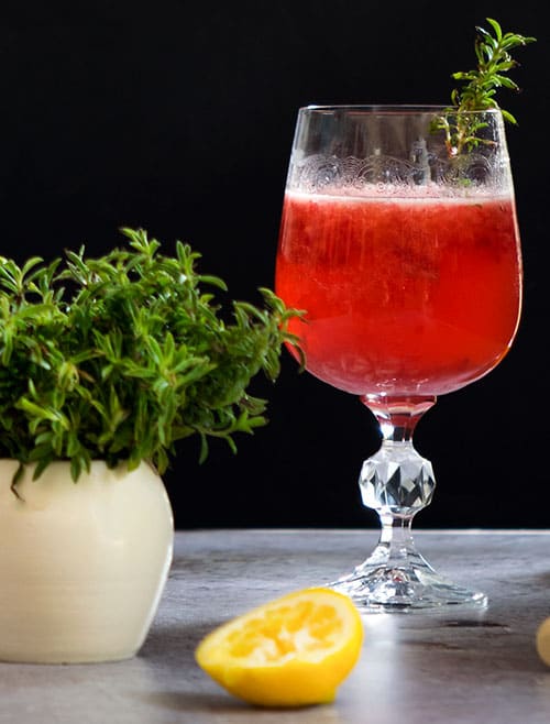 red fresh raspberry-thyme gin cocktail on a wine glass with 1/2 squeezed lemon in front, placed on a table, with black background