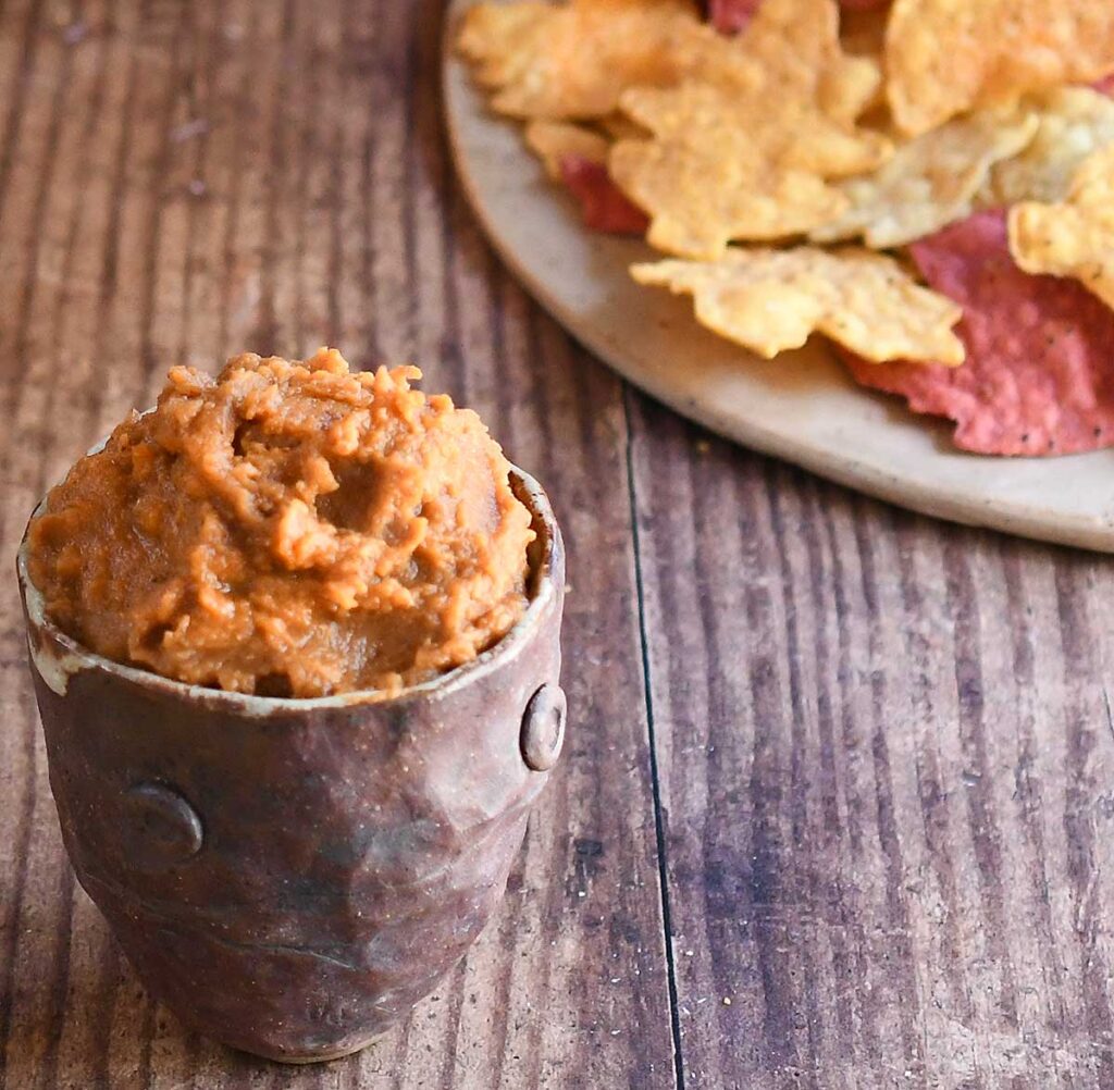 Pumpkin hummus in a brown potter bowl on a wooden table with leaf-shaped chips and a pie pumpkin.