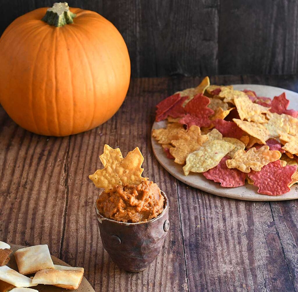 Pumpkin hummus in a brown potter bowl on a wooden table with leaf-shaped chips and a pie pumpkin.