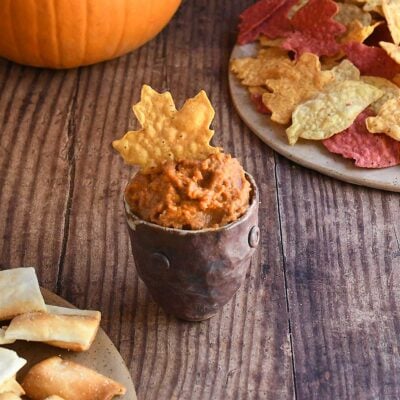 Ancho chile pumpkin hummus in a brown handforn potter bowl with a maple leaf shaped yellow tortilla chip