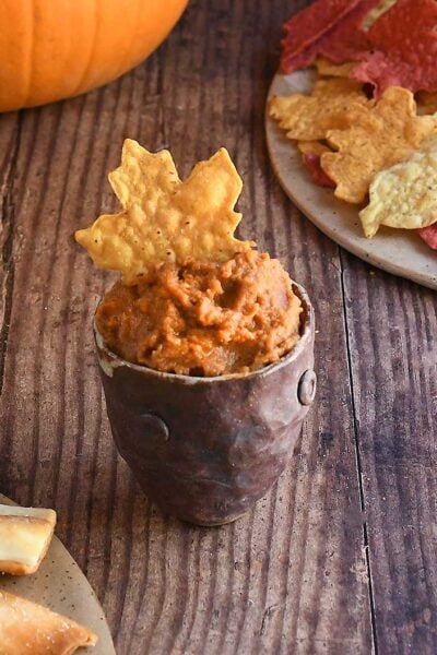 Ancho chile pumpkin hummus in a brown handforn potter bowl with a maple leaf shaped yellow tortilla chip