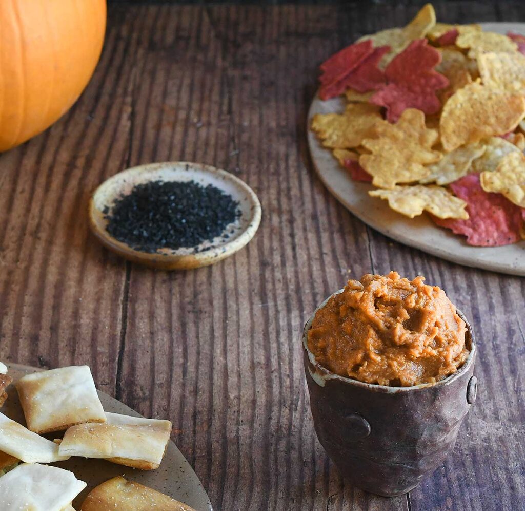 Pumpkin hummus in a brown potter bowl on a wooden table with leaf-shaped chips and a pie pumpkin.
