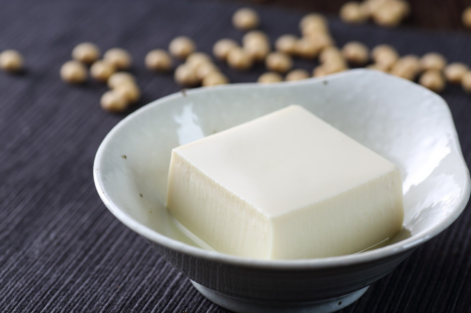 silken tofu on a small white bowl on top of a dark colored table with scattered soy beans in the background
