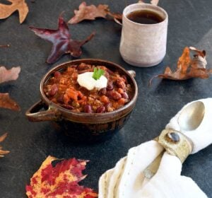 Vegan chili in a ceramic bowl with 2 handles, hot tea, on a slate table with autumn leaves.