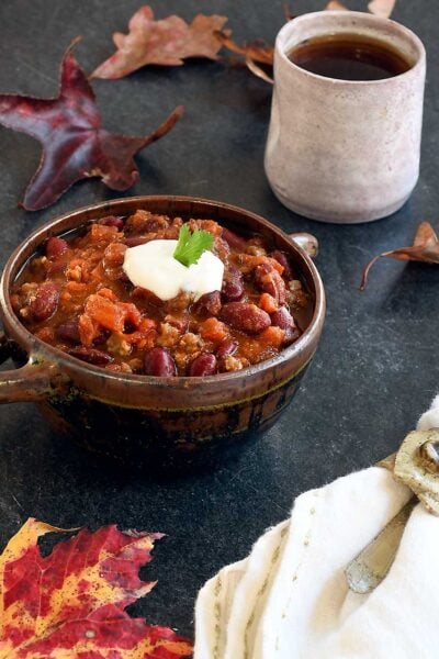 Vegan chili in a ceramic bowl with 2 handles, hot tea, on a slate table with autumn leaves.
