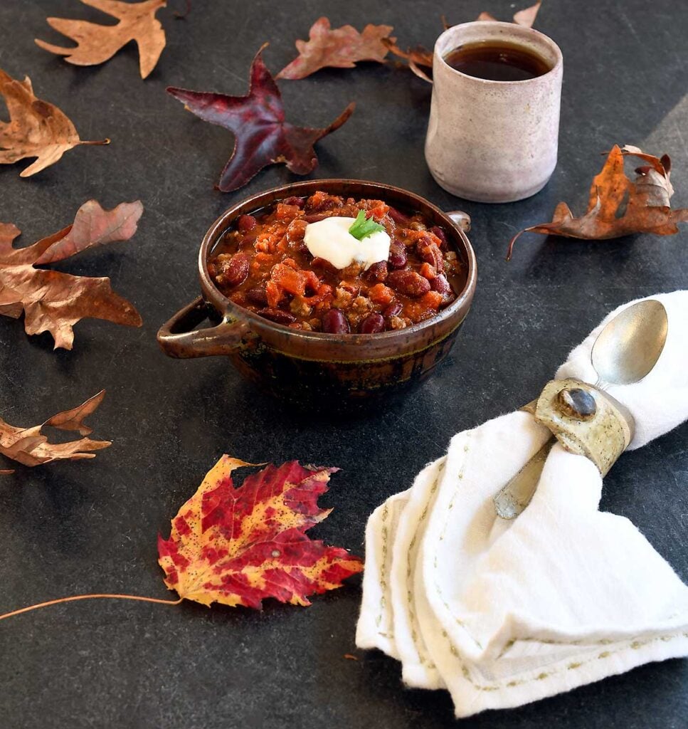 Vegan chili in a brown ceramic bowl with 2 handles, hot tea in a white ceramic mug, on a slate table with scattered autumn leaves.
