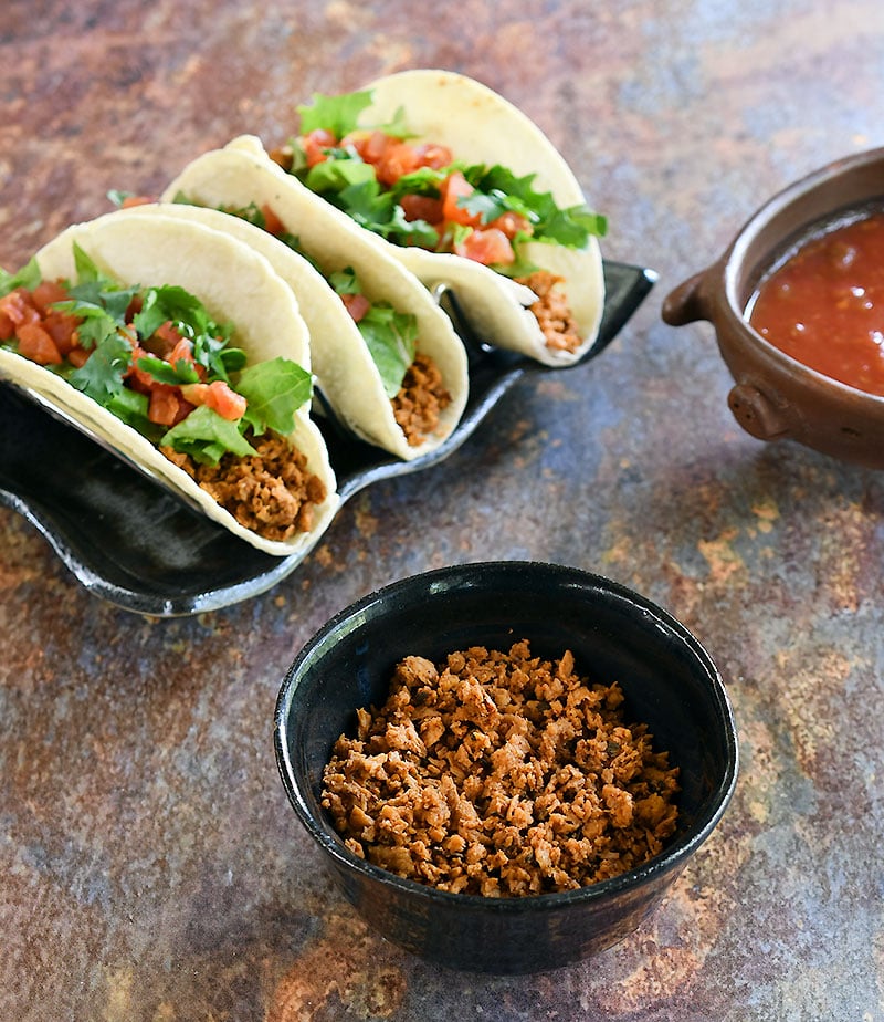 3 soft corn tacos on a black rectangle plate with red salsa in a pottery pig and soy curl taco crumbles in a small bowl.