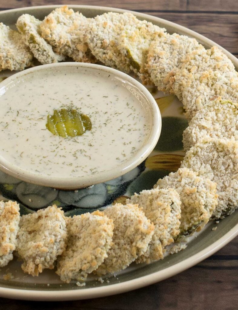 round serving platter with vegan air-fried pickle chips arranged in a ring on the outside edge with a bowl of vegan ranch dressing garnished with a pickle slice in the center.