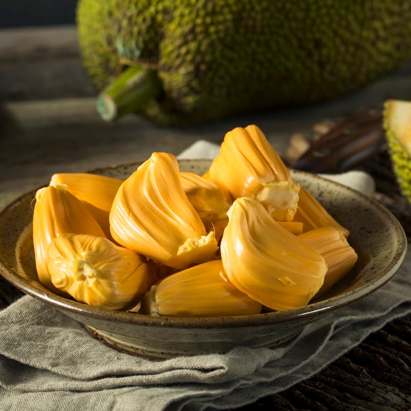 Jackfruit pieces in a plate sitting on a table with one whole jackfruit and a slice of it showing in the background