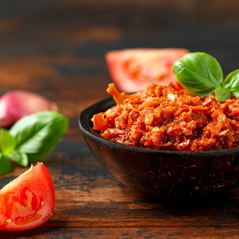 Sun dried tomatoes on a small black bowl sitting on a wooden chopping board with some fresh veggie cuts on the side